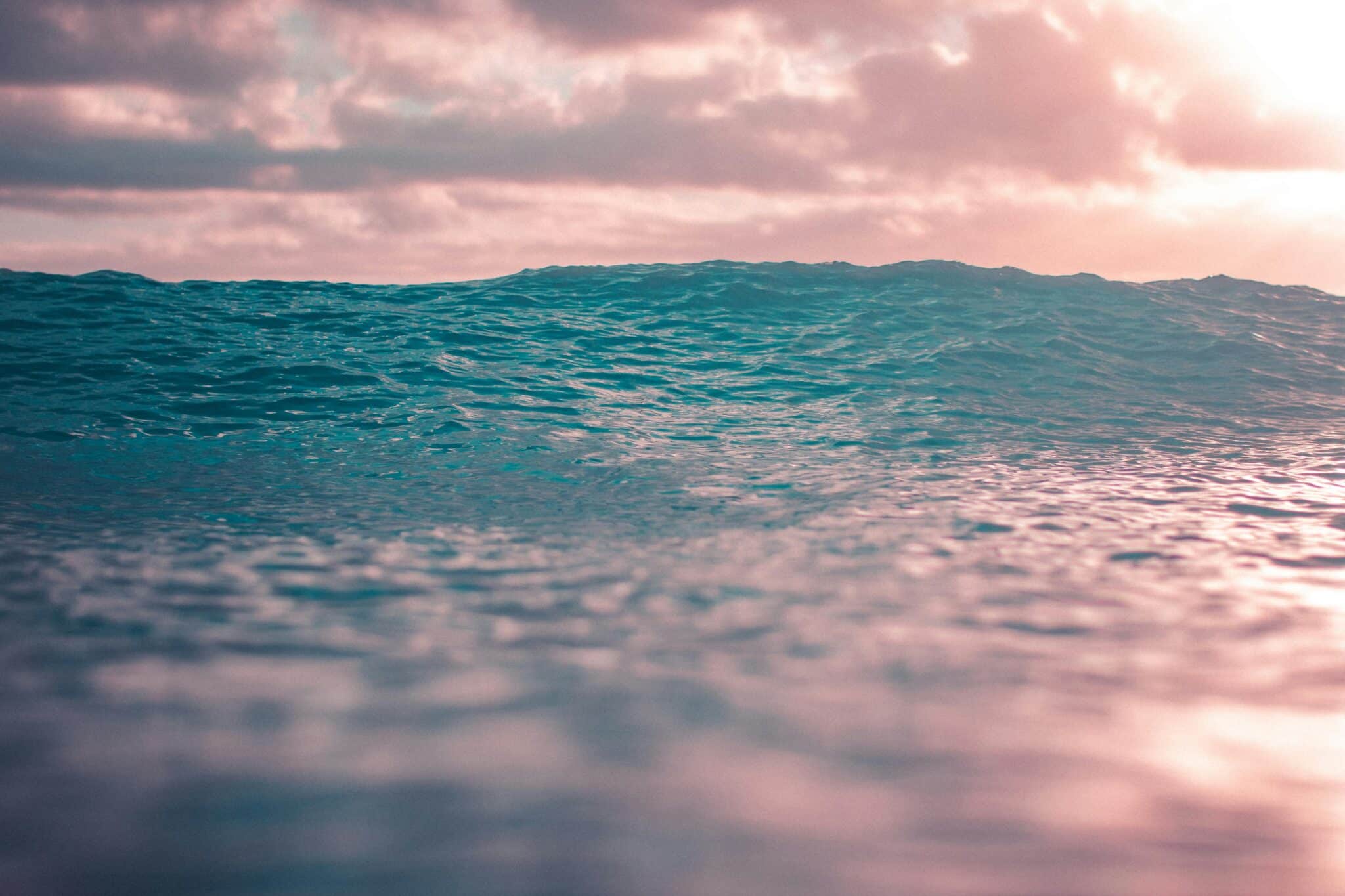 Meren aaltoilevaa pintaa ja pilvistä taivasta, jonka punerrus heijastuu mereen.