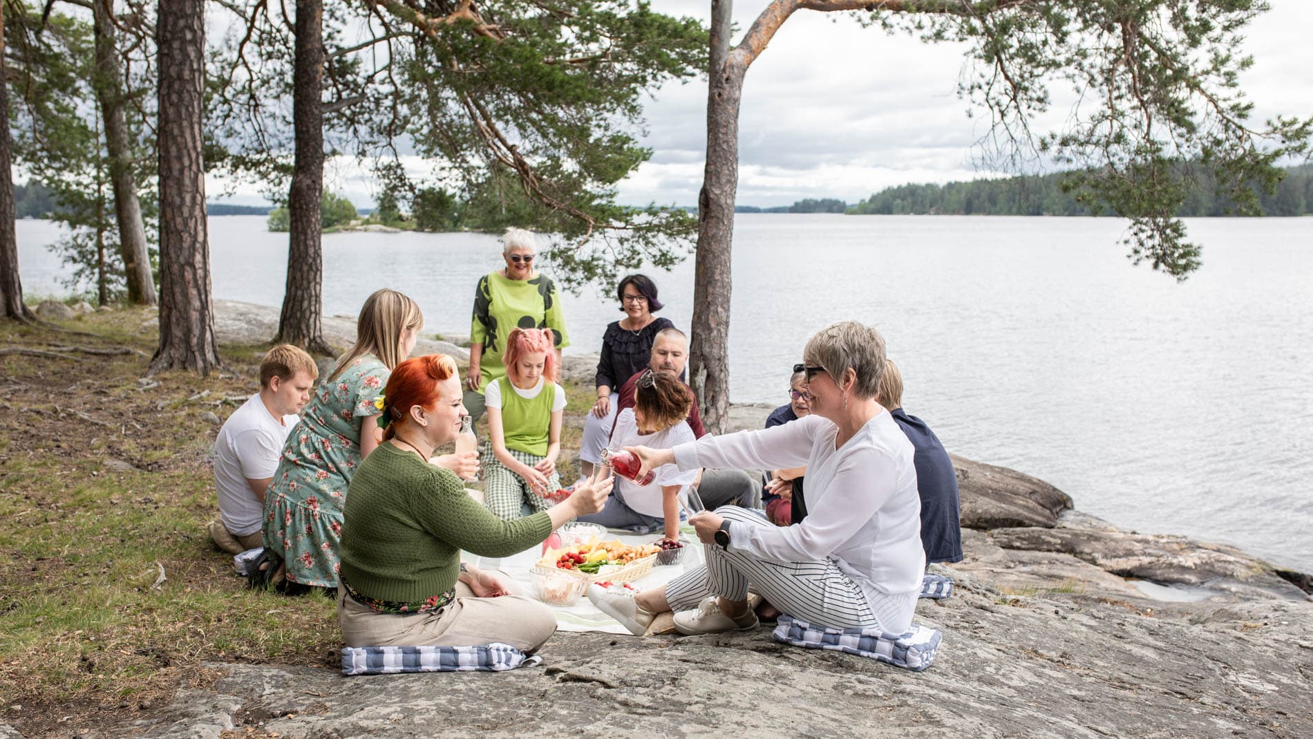 Joukko eri-ikäisiä naisia ja miehiä istuu piknikillä kalliolla järven rannalla. Joukon keskelle on levitetty kangas, jolle on katettu ruokaa.